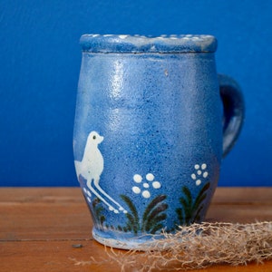 Può includere: Una tazza in ceramica blu con un uccello bianco e un motivo floreale. La tazza ha un manico e una forma leggermente arrotondata.