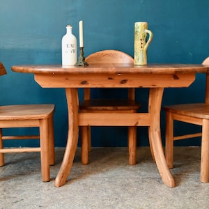 Peut inclure: Une table en bois avec une finition brun clair et un plateau ovale arrondi. Trois chaises avec une finition brun clair entourent la table. Une bouteille en céramique blanche avec du texte noir qui lit "Eau de Vie" est posée sur la table. Un vase en céramique vert avec un motif floral est posé sur la table.