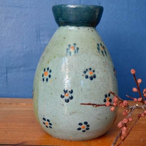 Peut inclure: Un petit vase en céramique bleu clair avec un bord bleu foncé et un motif floral orange et noir. Le vase est posé sur une surface en bois.