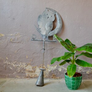 Op de afbeelding: Een metalen uil en halve maan sculptuur op een metalen paal met een groene plant in een pot op de voorgrond. De sculptuur is bevestigd aan een muur met een ruw, getextureerd oppervlak.