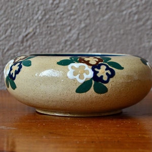 May include: A small, round, beige ceramic bowl with a floral design. The flowers are painted in shades of white, blue, brown, and green.