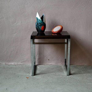 May include: A modern side table with a dark brown top and chrome legs. On the table are a decorative vase with blue, black, and red accents, and a small orange and white decorative object. The table is set against a pink wall.