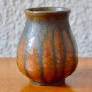 May include: A small, round ceramic vase with a brown and grey glaze. The glaze has a drip effect, creating vertical lines of grey on the brown background.
