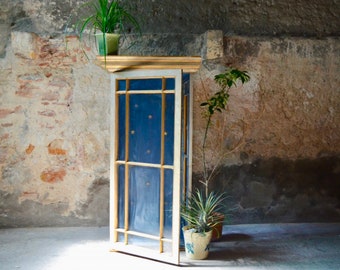 Op de afbeelding: Een hoge, witte kast met een blauwe deur en gouden versieringen. De deur heeft een rasterpatroon en staat licht open. De kast staat in een kamer met een bakstenen muur en planten.