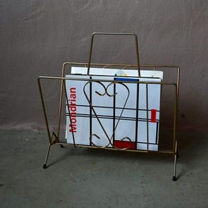 May include: A gold-toned metal magazine rack with a handle. The rack holds a magazine with a Mondrian-style design in red, white, black, and blue. The rack has a minimalist design with a heart-shaped detail.