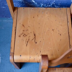 May include: Close-up of the top of a wooden chair with a light brown finish. The chair has a worn surface with scratches and marks.