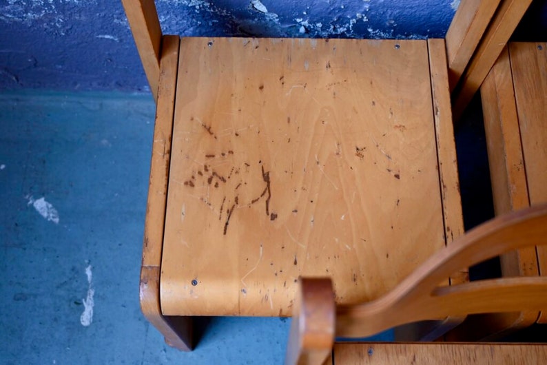 May include: Close-up of the top of a wooden chair with a light brown finish. The chair has a worn surface with scratches and marks.