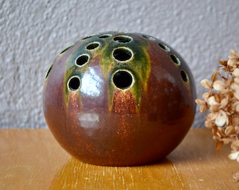 May include: A round, brown ceramic vase with green accents and holes for flowers. The vase has a glossy finish.