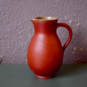 May include: A red ceramic jug with a rounded body and a handle. The jug has a light brown rim.