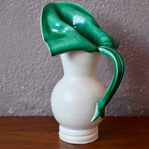 May include: A white ceramic jug with a green glazed handle and spout shaped like a flower. The jug is a decorative piece and is not intended for use.