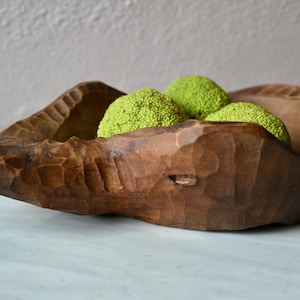Könnte beinhalten: Eine dunkelbraune, handgeschnitzte Holzschale mit rauer, facettierter Außenstruktur und einem länglichen, spitz zulaufenden Ende. Die Schale enthält drei leuchtend hellgrüne, texturierte, kugelförmige Objekte, die das Innere füllen.