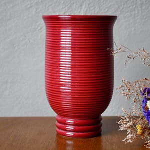 May include: A red ceramic vase with a ribbed design. The vase has a wide mouth and a narrow base.