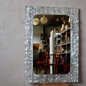 May include: Rectangular mirror with a textured, clear glass frame. The mirror reflects a room with shelves, chairs, and other objects. The frame has a unique, irregular surface.
