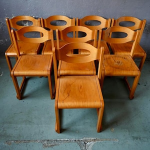 May include: A set of eight wooden chairs with curved backs and seats. The chairs are arranged in a row, with the backs facing the viewer. The chairs are made of a light brown wood and have a natural finish.
