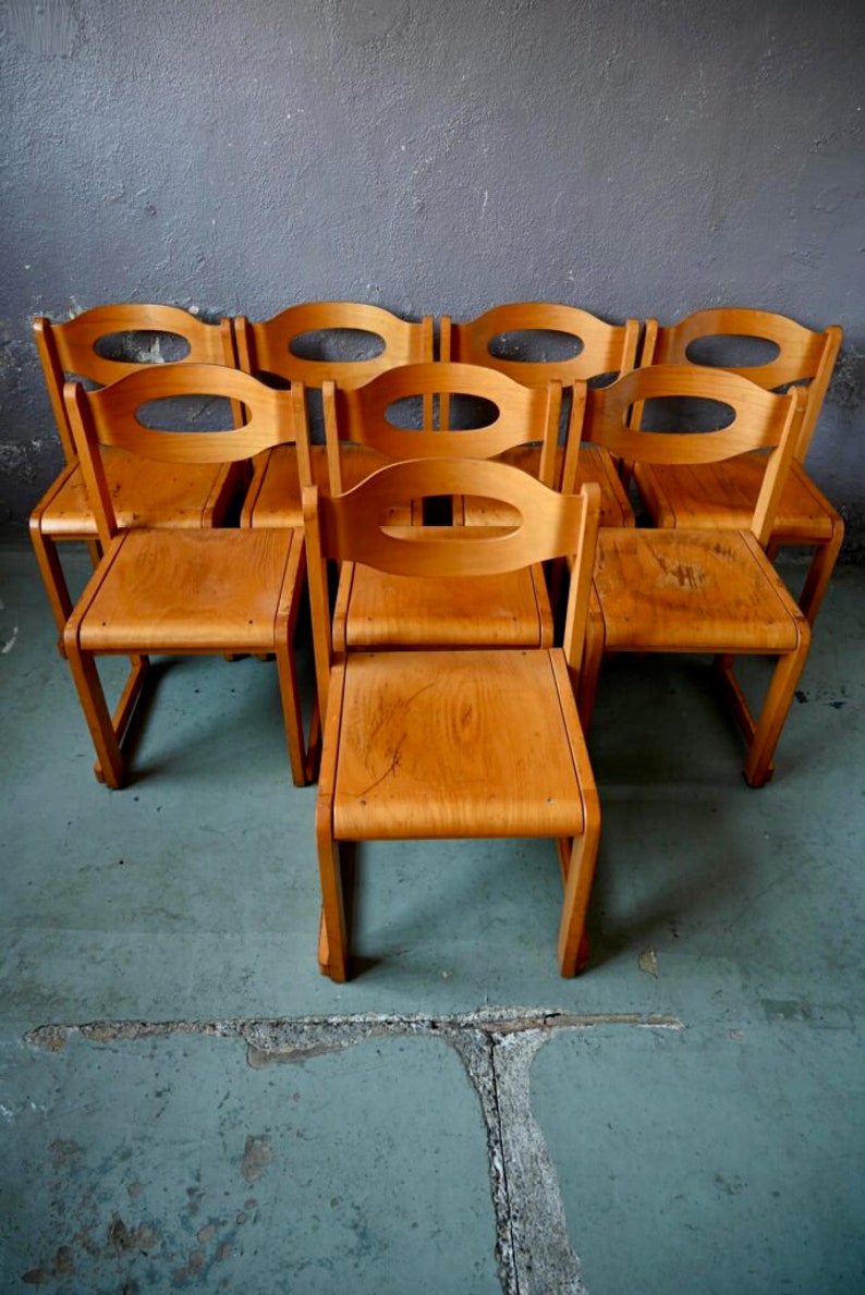 May include: A set of eight wooden chairs with curved backs and seats. The chairs are arranged in a row, with the backs facing the viewer. The chairs are made of a light brown wood and have a natural finish.