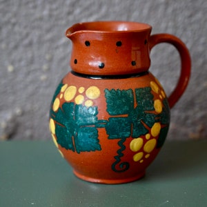 May include: A brown ceramic jug with a green and yellow floral design. The jug has a handle and a rounded body.