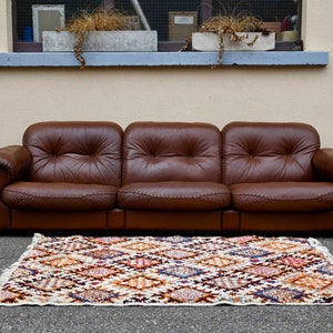 Peut inclure: Un canapé en cuir marron à trois places avec des coussins capitonnés. Le canapé est devant un mur blanc et une fenêtre avec des plantes en pots.