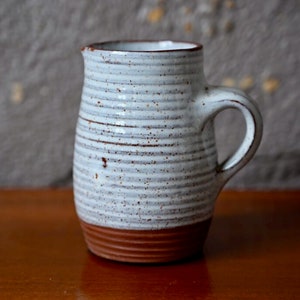 Puede incluir: Una jarra de cerámica blanca con un borde marrón y un esmalte moteado. La jarra tiene un cuerpo redondeado y un asa.