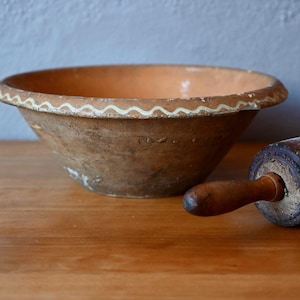 Puede incluir: Un cuenco rústico de cerámica con un diseño ondulado crema alrededor del borde. Un rodillo de madera con un mango oscuro descansa junto al cuenco. El cuenco y el rodillo están sobre una superficie de madera.