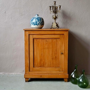 Peut inclure: Un meuble en bois clair à une porte, surmonté d'un vase à motifs bleus et blancs et d'un candélabre en laiton. Deux bouteilles en verre vert sont devant le meuble. Le meuble est en bois clair.