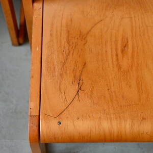 May include: A close-up of the wooden seat of a chair. The seat is made of light wood and has a few scratches on it.