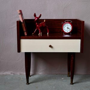 Può includere: Un comodino moderno della metà del secolo con finitura in legno scuro, un cassetto color crema e gambe sottili. Una statuina di cervo rosso, una sveglia rossa e un libro poggiano sopra. Il comodino è alto circa 61 cm.