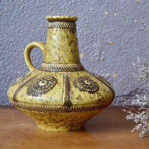 Peut inclure: Un vase en céramique jaune avec une surface texturée et des accents bruns. Le vase a une base large et un col étroit avec une poignée.