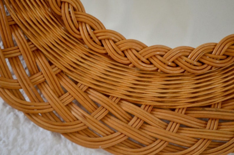 May include: Close-up of a woven wicker mirror frame. The frame is made of natural brown wicker and has a braided design around the edge.