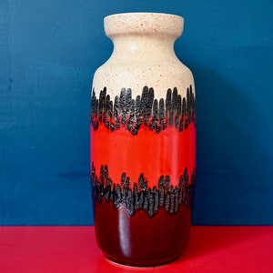 Peut inclure: Un vase en céramique avec un glaçage blanc, rouge et brun. Le vase a un motif noir en forme de gouttes autour du milieu.