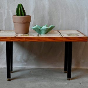 Peut inclure: Une petite table en bois avec un plateau en carrelage à motif floral. La table a des pieds en métal noir avec des accents dorés et est décorée d'un bol en céramique vert et d'un cactus en pot.