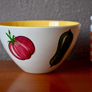 May include: A white ceramic bowl with a yellow rim and hand-painted illustrations of a tomato and a cucumber. The bowl is sitting on a wooden surface.