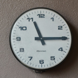 May include: A white analog clock with black hands and black numbers on a white face. The clock is in a black circular frame and has the word "Bodet" printed on the face.