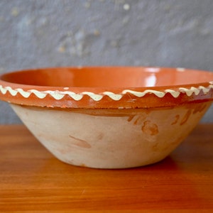 Puede incluir: Un cuenco rústico de terracota con un borde ancho y un patrón ondulado de color crema. El cuenco tiene un interior y exterior de color marrón anaranjado cálido, y está sobre una superficie de madera. Una pieza decorativa para la cocina.