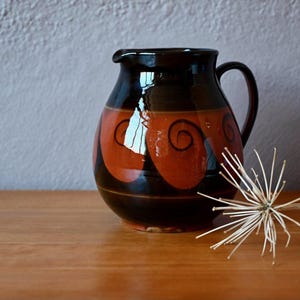Peut inclure: Une cruche en céramique avec un vernis brun foncé brillant présentant une large bande de couleur orange brûlé au centre. La section orange est décorée de motifs en spirale peints à la main en noir. La cruche possède une anse courbée.