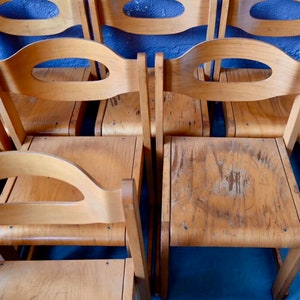 May include: A row of six wooden chairs with oval cutouts in the backrests. The chairs are made of light brown wood and have a worn, vintage look.