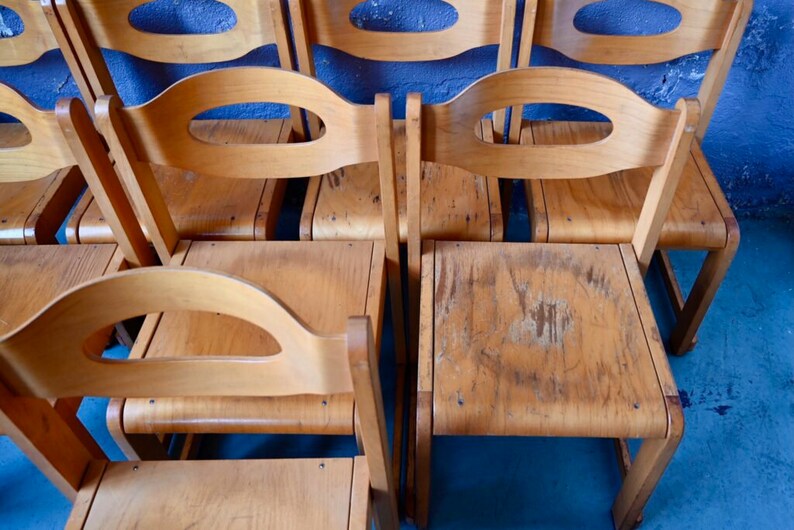 May include: A row of six wooden chairs with oval cutouts in the backrests. The chairs are made of light brown wood and have a worn, vintage look.