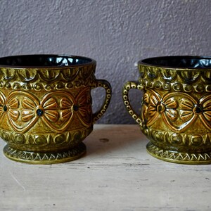 Peut inclure: Deux pots en céramique verts avec des motifs floraux marrons et des accents bleus. Les pots ont deux poignées de chaque côté.