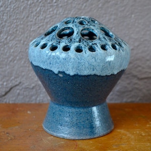 Puede incluir: Un jarrón de cerámica azul y blanco con una abertura redonda ancha y una base estrecha. El jarrón tiene un esmalte moteado y un anillo de agujeros alrededor de la parte superior.