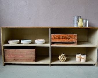 Op de afbeelding: Een houten rek met drie planken. De bovenste plank heeft een metalen container, een gele keramische kan en een metalen container. De middelste plank heeft een houten kist met het woord "Schweppes" erop gedrukt. De onderste plank heeft een houten kist met het woord "Tonka" erop gedrukt, een gele keramische kan met een geometrisch patroon en twee metalen containers.