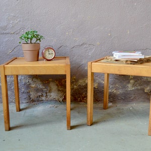 Puede incluir: Dos mesas auxiliares de madera con tapas cuadradas y cuatro patas. La mesa de la izquierda tiene una pequeña planta en maceta y un despertador rojo en la parte superior. La mesa de la derecha tiene una pila de libros en la parte superior.