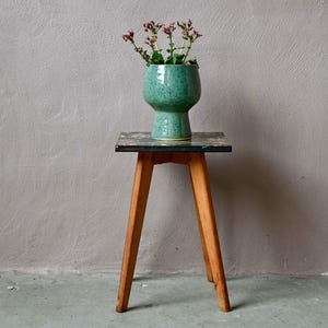 May include: A small wooden stool with four angled legs and a square top featuring a dark, patterned surface. Resting on the stool is a speckled, light green ceramic vase with a wide, rounded body and a narrow base, containing small pink flowers.
