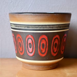 Può includere: Un vaso di ceramica con un motivo marrone, crema e nero. Presenta un motivo ripetuto di forme ovali rosse bordate di nero. La parte superiore e la base sono marrone chiaro.