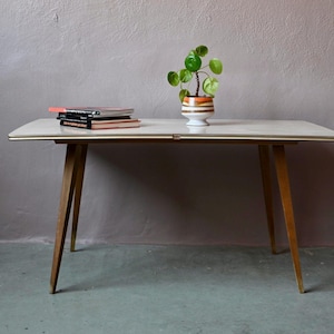 Può includere: Un tavolo bianco con una struttura in legno chiaro e quattro gambe. Il tavolo ha un piano bianco ed è decorato con una piccola pianta in vaso.