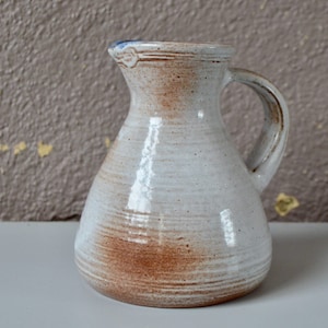 Puede incluir: Una jarra de cerámica blanca con un esmalte marrón y un borde azul. La jarra tiene un cuerpo redondeado y un asa.