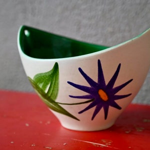 May include: A white ceramic bowl with a green rim and a painted design of a purple flower with a yellow centre and green leaves.