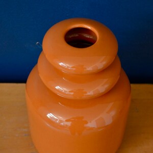 May include: A bright orange ceramic vase with a unique stacked design. The vase has a smooth, glossy finish and a wide opening at the top.