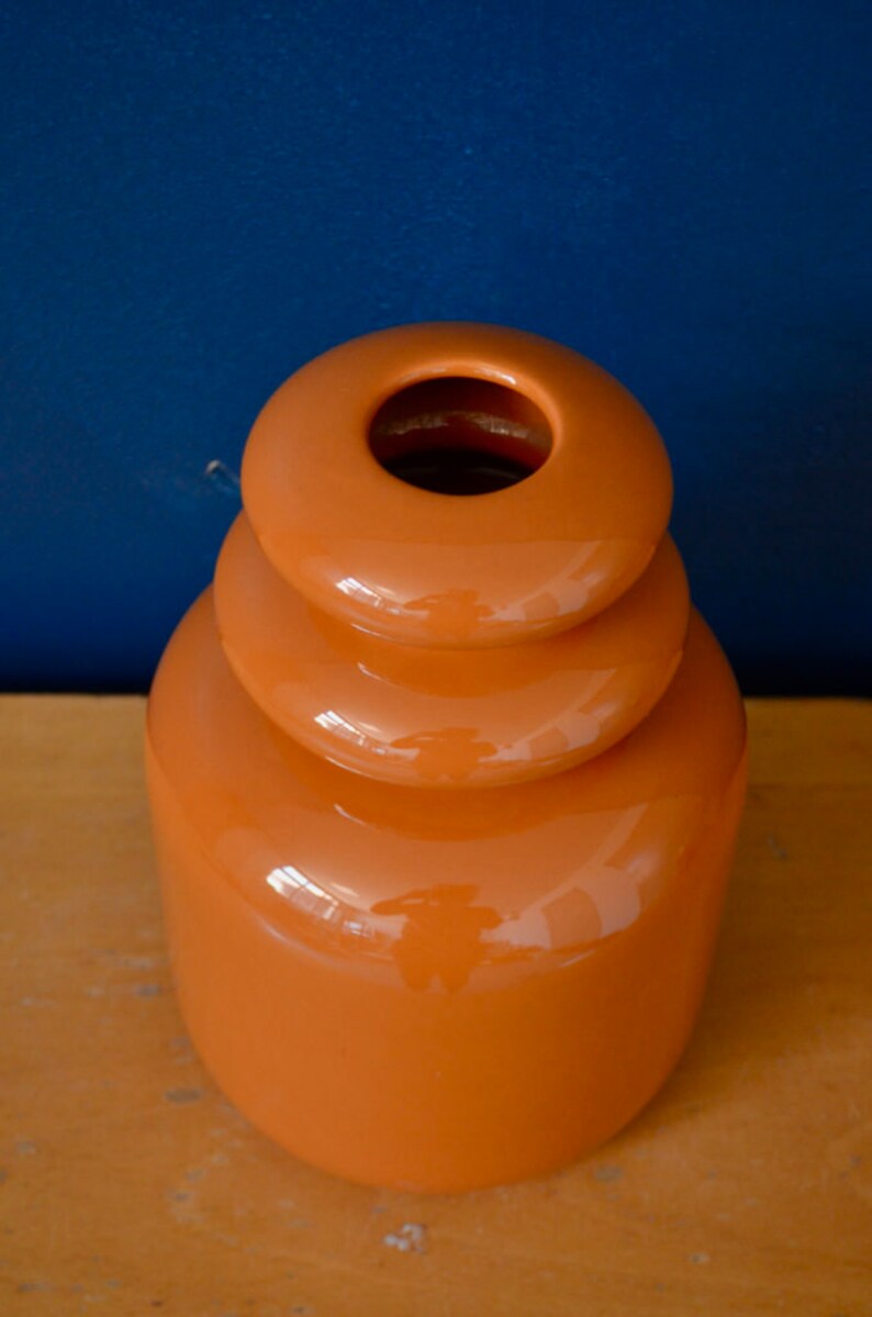 May include: A bright orange ceramic vase with a unique stacked design. The vase has a smooth, glossy finish and a wide opening at the top.