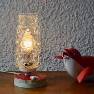 May include: A white and red table lamp with a clear bubble glass shade. The lamp is turned on and the light is shining. A red and white toy seal is sitting on the table next to the lamp.