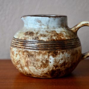 Puede incluir: Una jarra de cerámica marrón y blanca con cuerpo redondeado y asa. La jarra tiene un esmalte blanco con vetas y líneas marrones.