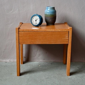 Può includere: Un piccolo comodino in legno con un cassetto, con una sveglia blu e un vaso decorativo. Il tavolo è in legno chiaro e ha quattro gambe. Il vaso è blu, marrone e verde.